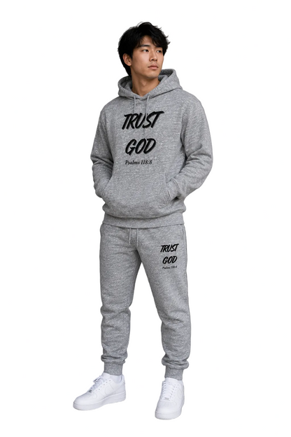 Person wearing a gray hoodie and jogger set with 'Trust God' text against a concrete wall.